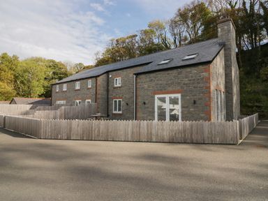 A stone cottage with multiple windows and a wooden fence around it surrounded by trees at Edgemere Cottage in Cartmel