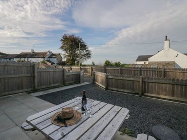 An outdoor wooden picnic table with a bottle and four glasses on a paved and gravel patio with a wooden fence and houses in the background at Edgemere Retreat in Cartmel