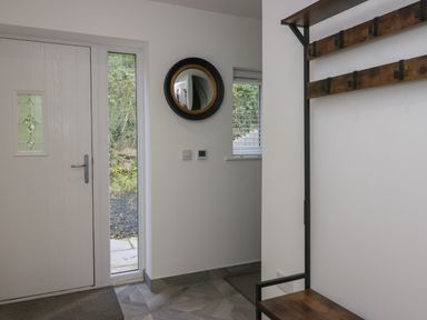 An entrance area with a white door a narrow window a round mirror on the wall and a wooden bench with coat hooks at Edgemere Retreat in Cartmel