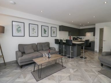 A living room with a gray sofa a wooden coffee table with wine glasses and a bottle stools at a kitchen island with dark cabinets in Edgemere Retreat Cartmel