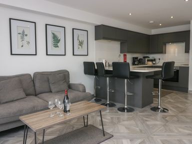 A living room with a gray sofa a wooden coffee table with a bottle and glasses and a kitchen with bar stools at Edgemere Retreat in Cartmel