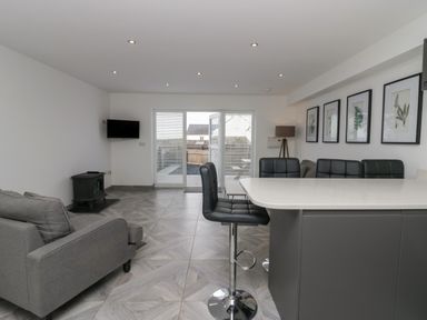 A living area with a gray sofa a small black stove a wall-mounted tv black bar stools a countertop and framed botanical prints at Edgemere Retreat in Cartmel