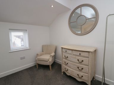 A small room with a beige armchair next to a wooden chest of drawers and a round mirror on the wall at Edgemere Retreat in Cartmel