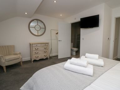 A bedroom with a bed, folded towels, an armchair, a chest of drawers, a round mirror, a wall-mounted television, and an open door leading to a bathroom at Edgemere Retreat in Cartmel