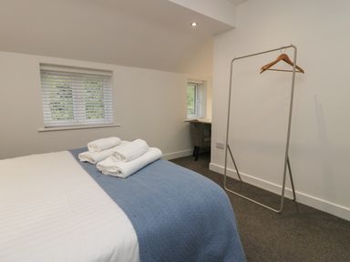 A bedroom with a bed covered by white and blue bedding with towels on top a clothes rack with hangers and a desk with a chair by a window at Edgemere Retreat in Cartmel
