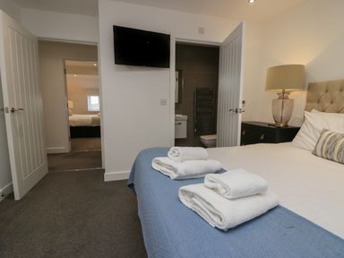 A bedroom with a bed with towels on it a bedside table with a lamp a wall-mounted TV and open doors showing a bathroom and another bedroom at Edgemere Retreat in Cartmel