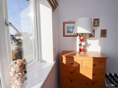 A window with a vase of seashells on the sill next to a wooden chest of drawers with a lamp and framed pictures on the wall at Coastal Retreat in Seahouses