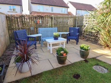 A backyard patio with blue and light blue wooden chairs a white table and several potted plants at Coastal Retreat in Seahouses
