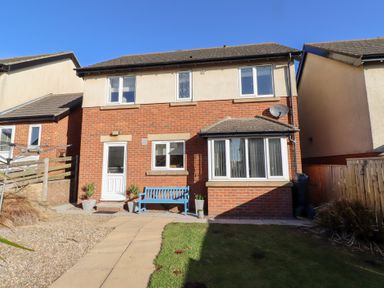 The back of a two story brick and stucco house with a white door blue bench and garden at Coastal Retreat in Seahouses