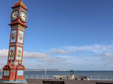 A clock tower next to the sea with benches along the pathway at Apartment 1 Weymouth