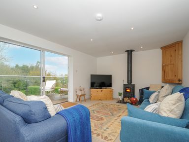 A living room with blue sofas a wood stove a television and glass doors leading to a balcony at Seamay in Polzeath