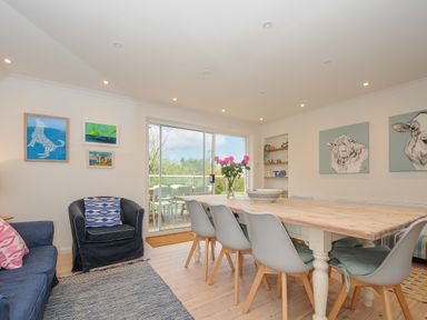 A dining room with a wooden table surrounded by chairs and a vase with flowers on it a living area with armchairs and artwork on the walls at Seamay in Polzeath