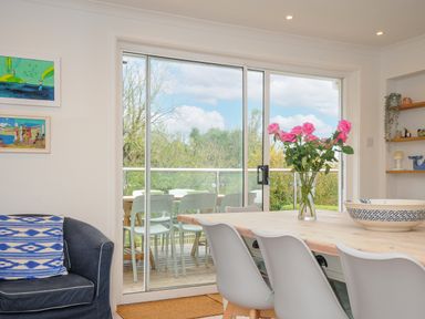 A dining area with a wooden table and white chairs a vase of pink flowers on the table and sliding glass doors leading to an outdoor seating area at Seamay in Polzeath