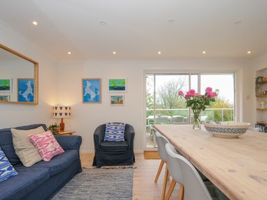 A living room with a blue sofa, an armchair, a wooden dining table with chairs and a vase of pink flowers at Seamay Polzeath