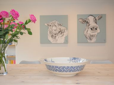 A wooden table with a blue and white ceramic bowl and pink roses in a glass vase with animal paintings on the wall at Seamay in Polzeath