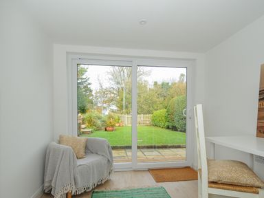 A room with a chair covered with a gray blanket and pillow a white desk with chairs and sliding glass doors leading to a garden at Seamay Polzeath
