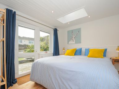 A bedroom with a bed blue and yellow pillows a painting of a rabbit bedside tables and glass doors leading outside at Seamay in Polzeath