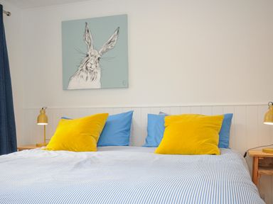 A bedroom with a bed with yellow and blue pillows and a rabbit painting above at Seamay Polzeath