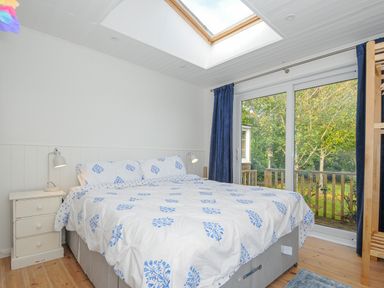 A bedroom with a double bed blue and white bedding bedside tables lamps a skylight and glass sliding doors with blue curtains overlooking trees at Seamay in Polzeath