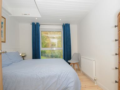 A bedroom with a bed covered in striped bedding a window with blue curtains a gray chair and a wooden chest of drawers at Seamay Polzeath