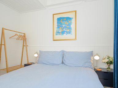 A bedroom with a bed with striped bedding two bedside tables with lamps flowers and a wooden clothes rack at Seamay in Polzeath