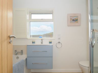 A bathroom with a blue vanity unit under a window a bathtub with a blue towel and a toilet at Seamay in Polzeath