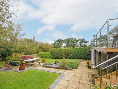 A garden with a picnic table a fire pit a small pond and a raised deck with glass railing at Seamay in Polzeath