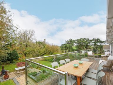 An outdoor deck with a wooden table surrounded by chairs and lounge chairs with cushions overlooking a garden at Seamay in Polzeath