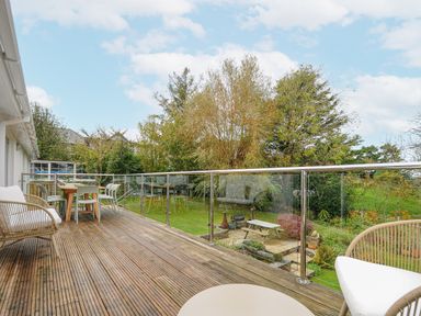 An outdoor wooden deck with chairs and a table overlooking a garden with trees and a picnic bench at Seamay in Polzeath