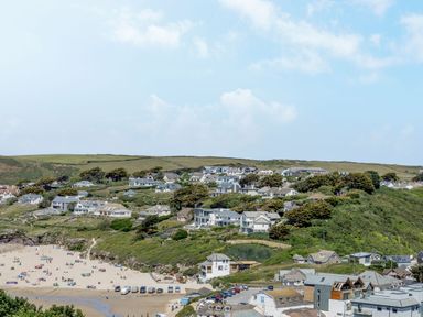 A coastal village with houses on a hill overlooking a beach with people and parked cars at Seamay in Polzeath