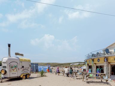 A beach area with a wood fired pizza trailer picnic tables and a surf hire shop at Seamay Polzeath