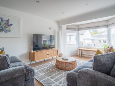 A living room with two grey sofas a wooden TV stand with a flat screen TV a round rattan coffee table a large window with bench seating and a colorful giraffe painting on the wall at Hoheck in Abersoch