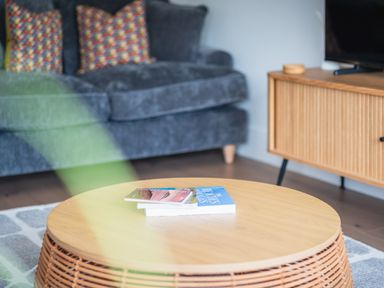 A round wooden coffee table with books in front of a grey sofa with patterned cushions and a wooden cabinet with a television at Hoheck in Abersoch