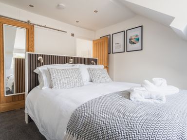 A bedroom with a bed with white linens and patterned pillows towels on the bed a wooden door with a mirror on the wall with framed pictures at Hoheck in Abersoch