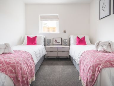 A bedroom with two single beds pink patterned throws and cushions a small cabinet with lamps and a picture on it at Hoheck in Abersoch