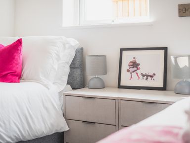A bedroom with a bed featuring white pillows and pink cushions next to a bedside table with two lamps and a framed picture at Hoheck in Abersoch