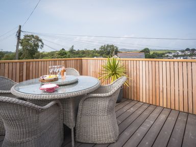 An outdoor patio with a glass top table and wicker chairs a bottle with four glasses and a wooden tray with food on the table and a potted plant against a wooden fence at Hoheck in Abersoch