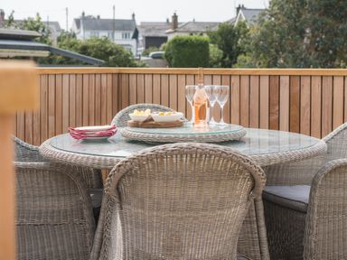 An outdoor wicker dining table with glass top set with wine glasses a bottle and bowls of snacks on a wooden deck at Hoheck in Abersoch