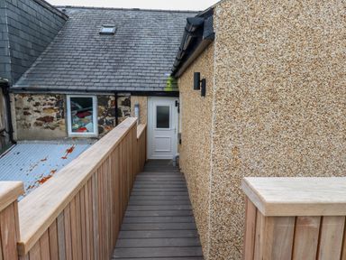 A wooden walkway leading to a white door on a building with a textured exterior wall and a slate roof at Hoheck in Abersoch