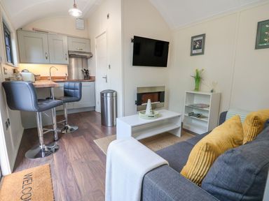 A compact living area with a gray sofa and yellow pillows a white coffee table a wall-mounted tv and a small kitchen with two bar stools at The Retreat in Bangor-On-Dee