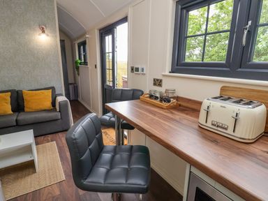 A kitchen and living area with a wooden counter, black chairs, a toaster, jars, a grey sofa with yellow cushions, and a white table at The Retreat in Bangor-On-Dee