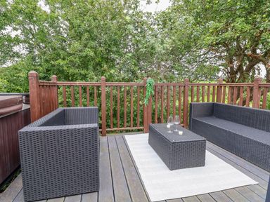 An outdoor patio with two black wicker sofas a matching table with glasses and candles on a white rug and wooden railing with trees behind at The Retreat in Bangor-On-Dee