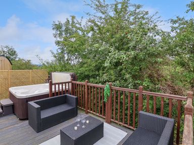 An outdoor wooden deck with a hot tub two black wicker sofas a black wicker coffee table with glasses and candles and trees in the background at The Retreat in Bangor-On-Dee