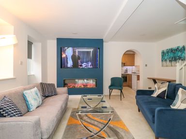 A living room with two sofas a glass coffee table a TV mounted above a fireplace and a view into a kitchen at 4 Noel Court in Chipping Campden
