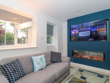 A living room with a grey sofa with three cushions, a glass coffee table, a built-in electric fireplace, and a mounted TV on a blue accent wall at 4 Noel Court in Chipping Campden