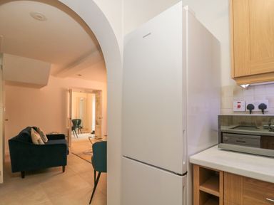 A kitchen area with a white Samsung refrigerator next to wooden cabinets and a microwave oven with a view into a living room with blue chairs and a sofa at 4 Noel Court in Chipping Campden