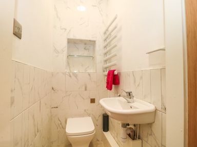 A small bathroom with a toilet, a wall-mounted sink, a towel rail with a red towel, and white marble tiles on the walls at 4 Noel Court in Chipping Campden