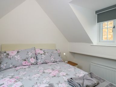 A bedroom with a bed with floral bedding and pillows a bedside table with a small lamp and a window with a gray blind at 4 Noel Court in Chipping Campden
