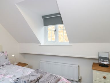 A bedroom with a bed with floral bedding a small window with a blind and a radiator below at 4 Noel Court in Chipping Campden