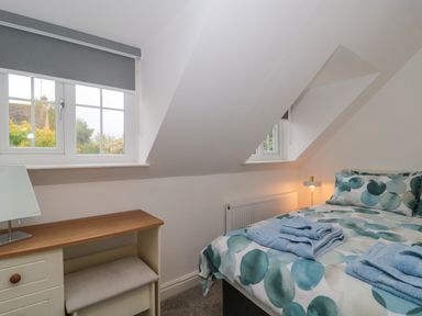 A bedroom with a bed, folded towels on the bed, a window with a grey roller blind, a small desk with a lamp, and a bench at 4 Noel Court in Chipping Campden